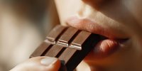 Close-up of person biting into a square of dark chocolate bar, showing sensual lips and fingers holding the treat. Perfect for food advertising.