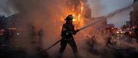 Firefighter in protective gear battles intense house fire with water hose as flames engulf residential building in dramatic emergency response scene.