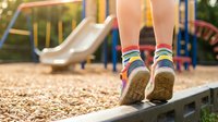 Child wearing colorful striped socks and sneakers walking on balance beam at playground with slide and equipment in blurred background during outdoor play.