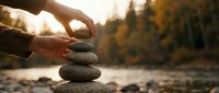 Hands carefully stacking smooth river stones in balanced tower outdoors during golden hour sunset with autumn forest background for meditation and mindfulness.