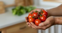 Fresh ripe cherry tomatoes on vine held in hands over kitchen counter with fresh produce and cutting board in background.