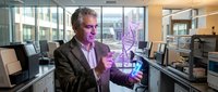Middle-aged Caucasian businessman in suit jacket presenting holographic DNA helix model in modern office laboratory with computers and scientific equipment.