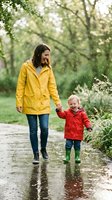 Young Caucasian mother in yellow raincoat walks with laughing toddler in red jacket through rain puddles, holding hands outdoors.