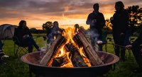 Group of friends gathering around campfire at sunset enjoying outdoor camping adventure with warm flames and golden sky creating cozy atmosphere for bonding.