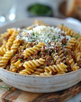 Rotini pasta with hearty meat sauce topped with grated parmesan cheese and fresh herbs served in rustic white ceramic bowl, close-up view of traditional Italian meal.