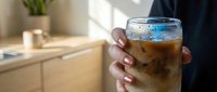 Person holding glass of cola with ice cubes in modern kitchen interior with natural light and wooden furniture.