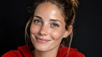 Young woman with freckles and natural beauty smiling warmly against black background, wearing bright red top, hair casually styled in updo.