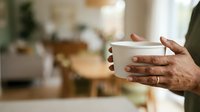 Close-up of hands holding white ceramic mug with warm beverage in modern kitchen setting, suitable for lifestyle, hospitality and beverage marketing campaigns.