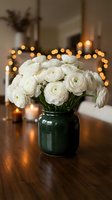 White ranunculus flowers in green ceramic vase on wooden table with warm bokeh lights creating cozy home interior atmosphere for spring decor and celebrations.