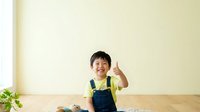 Young Asian boy wearing yellow shirt and denim overalls giving thumbs up gesture indoors. Isolated on light background, ready for design use on promotional materials and educational content.