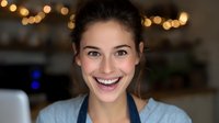 Young Caucasian woman with natural makeup and bright genuine smile wearing casual blue top against blurred bokeh lights background in cozy home setting. Positive emotional portrait.