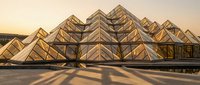Modern architectural structure with geometric glass and metal framework casting dramatic shadows during golden hour sunset, suitable for corporate, design, and contemporary building projects.