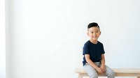 Young Asian boy, approximately 8-10 years old, wearing dark navy t-shirt and light gray pants, sitting on wooden bench against white wall background, smiling at camera.