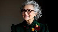 Senior woman with silver hair wearing glasses and emerald green dress with floral decorations smiles warmly against dark background, radiating elegance and confidence.
