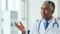 Mature African American male doctor in white coat with stethoscope gesturing while speaking in modern medical facility for healthcare presentations.