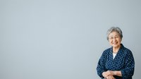 Senior woman with gray hair wearing navy blue patterned blouse, smiling at camera against neutral background. Isolated portrait for corporate, healthcare, lifestyle and editorial design use.