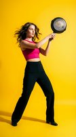 Young Caucasian woman in pink sports bra and black pants holding frying pan in dynamic pose on bright yellow background for fitness and cooking concepts.