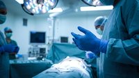 Surgeon putting on blue medical gloves in modern operating room with surgical lights and medical team preparing for procedure in hospital setting.