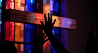 Silhouette of hands reaching toward illuminated stained glass cross in dark church interior creating spiritual worship atmosphere for religious content.