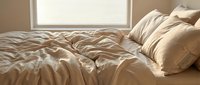 Unmade bed with beige linen sheets and pillows near sunlit window, conveying morning comfort and relaxation for bedroom design and lifestyle content.