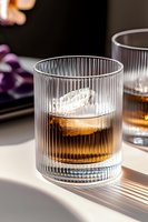 Whiskey with large ice sphere in ribbed crystal glass casting dramatic shadows on white surface, luxury cocktail photography in natural light.