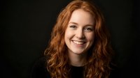 Young Caucasian woman with curly red hair smiling warmly at camera against dark background. Professional headshot for business, marketing materials.