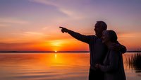 Father and son silhouettes pointing at sunset over calm lake water with orange purple sky reflection creating peaceful family bonding moment outdoors.