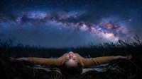 Young woman lying in grass field under spectacular Milky Way galaxy starry night sky, arms outstretched in peaceful contemplation and wonder.