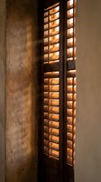 Warm golden light filters through wooden shutters creating dramatic shadows on textured concrete wall in modern interior design space.
