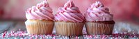 Pink frosted cupcakes with sprinkles arranged in row on rustic wooden surface against soft pink bokeh background, fresh homemade vanilla dessert treats.