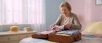Senior woman with blonde hair and fair complexion, wearing a cardigan, carefully folding colorful clothing into an open vintage suitcase on a bed in a bright bedroom with pastel walls and natural light from windows.
