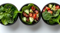 Fresh spinach salads with tomatoes, cucumbers and sesame seeds served in black bowls on white background, top view. Healthy vegetarian lunch concept.