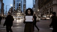 Young African American woman holding blank white sign on busy city street at dusk with illuminated skyscrapers and blurred pedestrians for protest or advertising mockup.