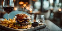 Gourmet burger with sesame bun, beef patty, fresh vegetables and golden fries served on wooden board in upscale restaurant with wine glass and warm bokeh lighting.