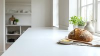 Freshly baked artisan bread loaf on white kitchen counter with natural window light, green plant, and modern minimalist interior design background.
