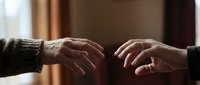Two hands reaching toward each other in a gesture of connection, support, or agreement. Warm indoor lighting creates an intimate atmosphere suitable for concepts of trust, partnership, help, and human connection.