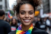 Young woman with dark curly hair and warm brown skin smiling at camera wearing rainbow pride flag colors at outdoor event celebration.