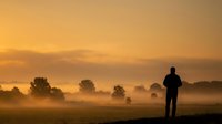 Man silhouette standing in misty field during golden sunrise with fog rolling over countryside landscape creating peaceful morning atmosphere for meditation.