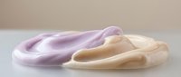 Soft pastel-colored slime or putty in purple and beige tones stretches and flows across a neutral surface, showcasing malleable texture for sensory play or stress relief.