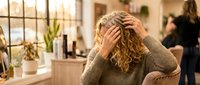 Stressed young blonde woman with curly hair holding head in hands showing frustration and overwhelm in modern office workspace environment.