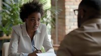 Young African American businesswoman in white blazer listening attentively during meeting, taking notes while discussing with colleague in modern office setting.