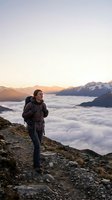 Male hiker with backpack standing on mountain trail at sunrise above clouds, surrounded by snow-capped peaks. Adventure and outdoor exploration concept for travel and lifestyle use.