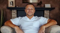 Confident mixed race male in white t-shirt relaxing on couch at home, displaying genuine smile and positive attitude. Modern living room interior with brick wall and decor.