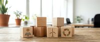 Wooden blocks with sustainability symbols including sun, tree, and recycling icon arranged on desk with potted plants and bright window light, representing eco-friendly business practices.