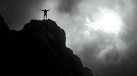 Silhouette of person standing with raised arms on mountain peak against dramatic stormy sky and sunburst through dark clouds. Concept of achievement and freedom.