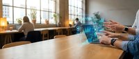 Businessman using holographic digital technology interface at modern office desk with colleagues working in background during daytime meeting.