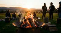 Group of friends gathering around crackling campfire at sunset in rural countryside with mountains in background for outdoor adventure and social bonding.