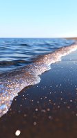 Gentle waves washing onto sandy beach with sparkling water droplets catching sunlight, creating magical bokeh effect along shoreline against blue ocean horizon.