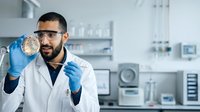 Young male scientist with dark hair and beard, Middle Eastern ethnicity, wearing white lab coat and blue gloves, holding petri dish in modern laboratory setting for research and development.