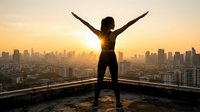 Woman silhouette with raised arms celebrating success on rooftop overlooking city skyline at golden hour sunrise creating motivational achievement moment.